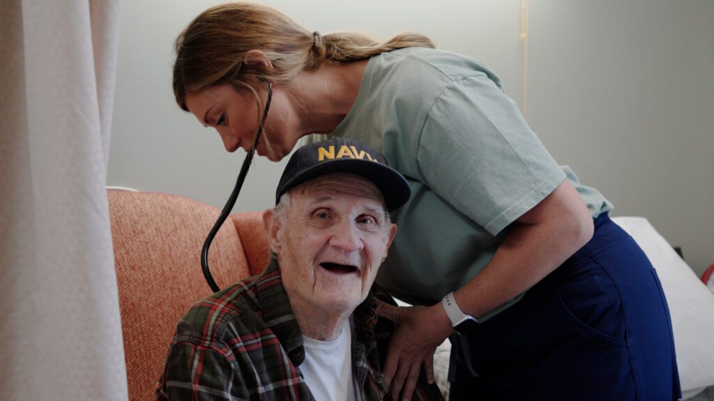 A man smiles while a nurse assists him at a skilled nursing facility